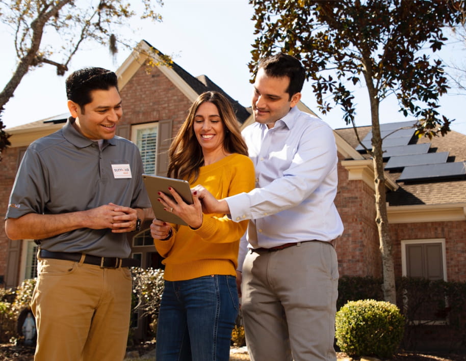 Three people standing in front of a house, examining a tablet.